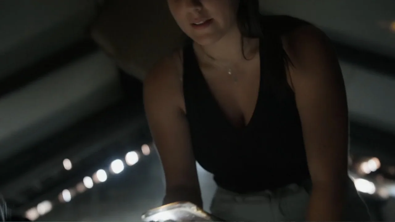 The Children of October 7 (2025) movie still: medium — A dimly lit indoor space with blurred bokeh lights in the dark background. A young woman in a…