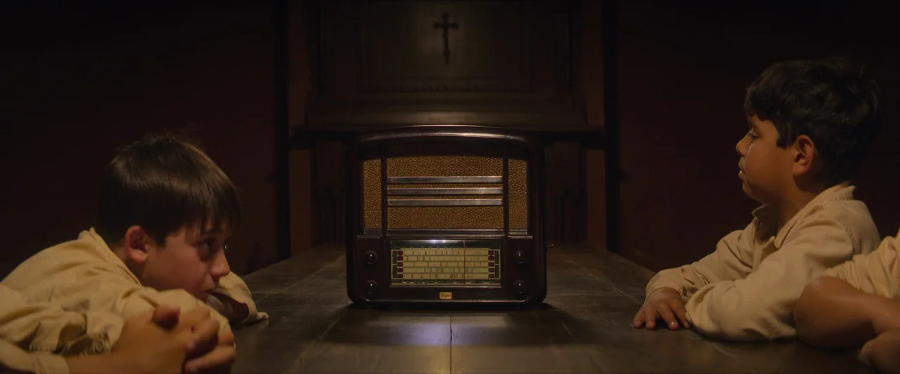 The New Boy (2023) movie still: medium — Two boys sit at a wooden table in a dimly lit, dark space. A vintage radio sits between them, casting a…