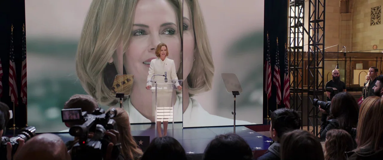 Long Shot (2019) movie still: fullbody — A woman stands on a blue stage featuring several American flags and large LED screens. A large projected…