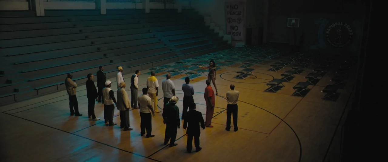 Chi-raq (2015) movie still: wide — An empty basketball court is used as a makeshift shelter with rows of cots. A group of men stands in a semicircle…