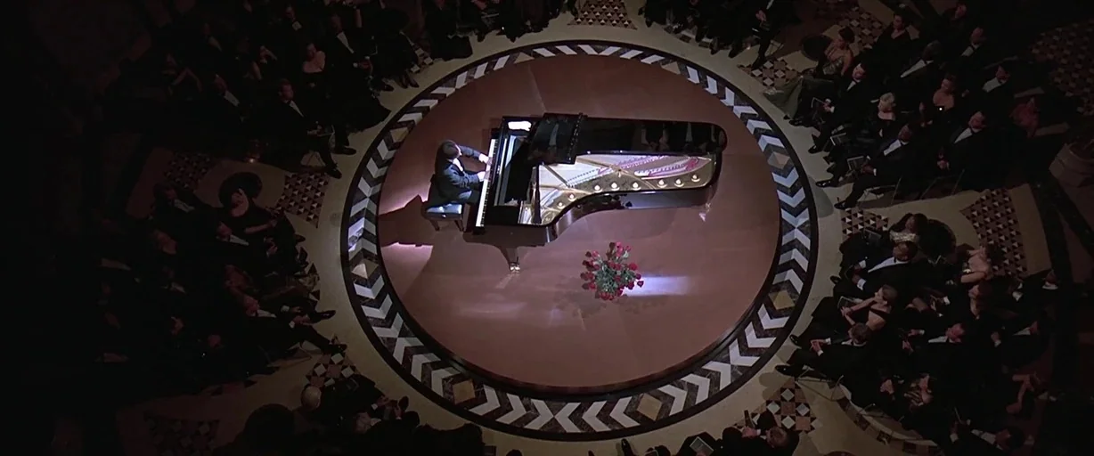 Gattaca (1997) movie still: wide — An elegant circular rotunda features a grand piano on a raised wooden stage. A lone man sits at the piano…