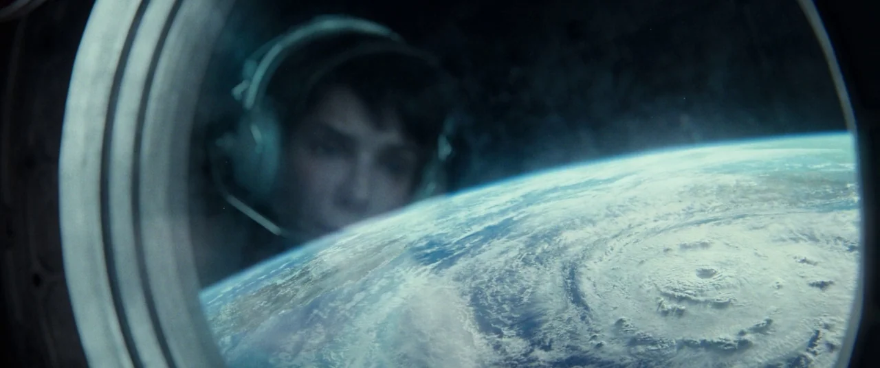 Gravity (2013) movie still: closeup — A view through a circular porthole reveals the curvature of the blue planet Earth. A female astronaut wearing…