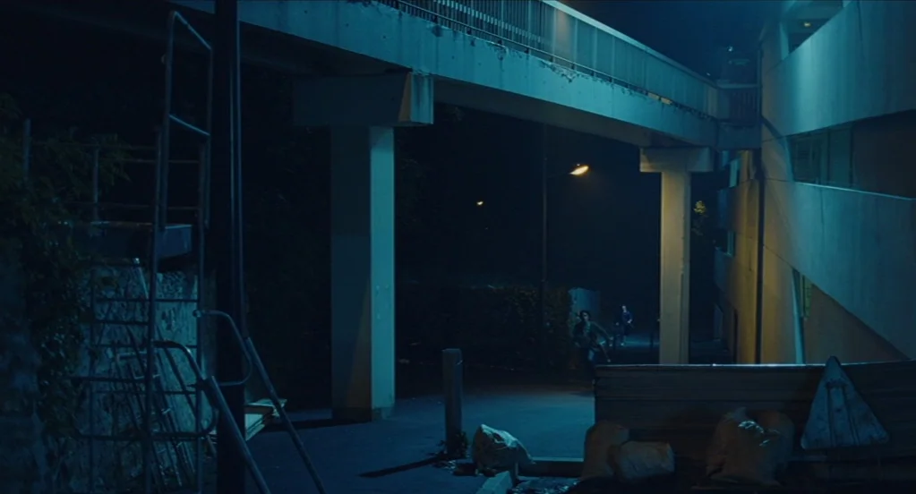 Something in the Air (2012) movie still: wide — A concrete pedestrian bridge spans across a darkened path at night. Several bags are piled on the…