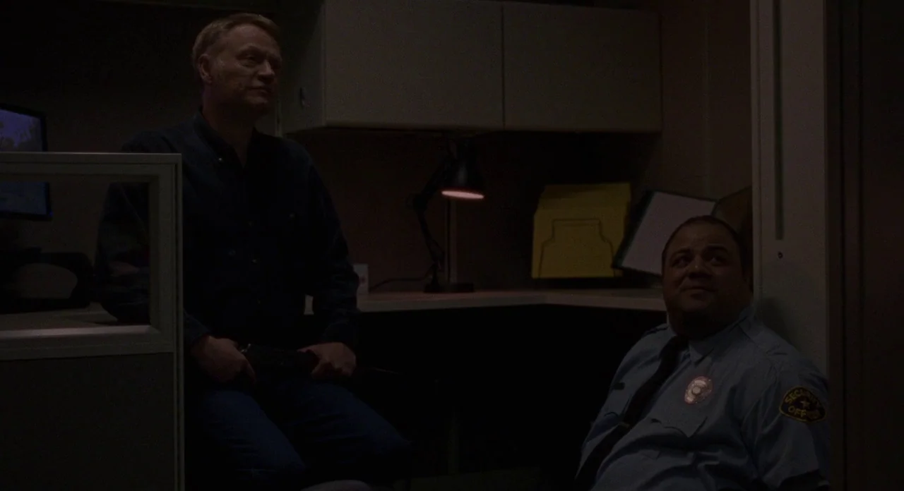 Certain Women (2016) movie still: medium — A dimly lit security station features a desk, filing cabinets, and office equipment. Two men sit across…