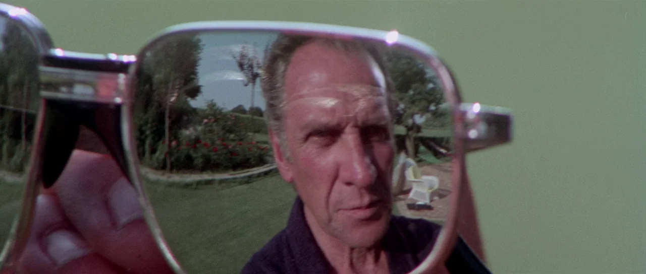 The Suspicious Death of a Minor (1975) movie still: closeup — A man's face is reflected in the lens of a pair of held eyeglasses. The background…