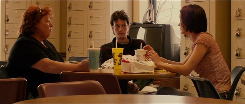 Bubble (2005) movie still: medium — A staff breakroom with lockers lining the back wall and plastic chairs at tables. Three people sit around a…