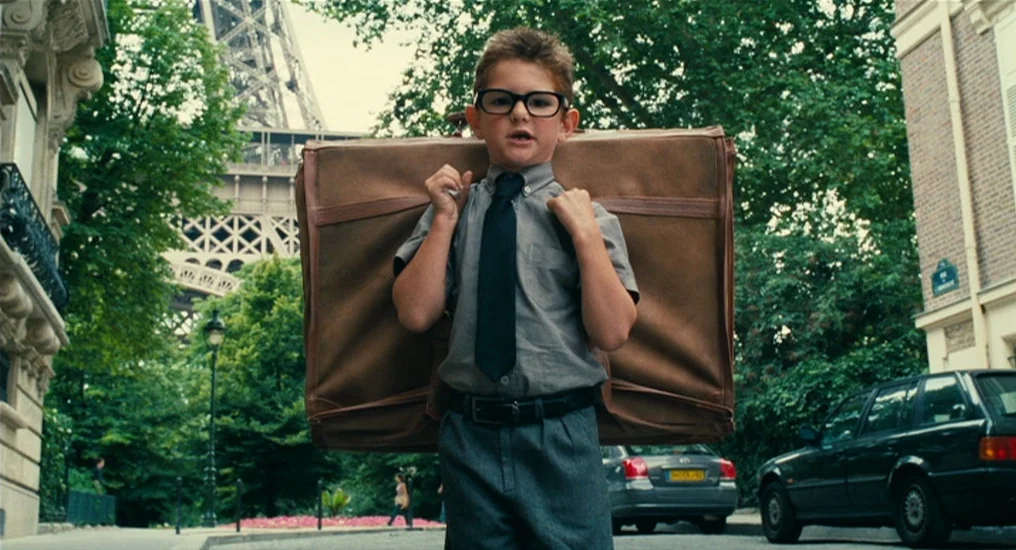 Paris, Je T’Aime (2006) movie still: medium — A young boy stands in a Paris street with the Eiffel Tower in background. He carries a large brown…