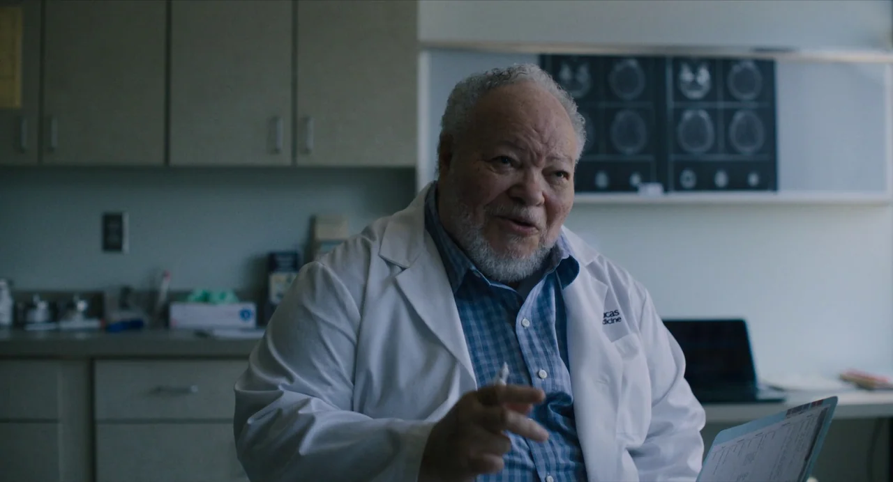 Causeway movie still: medium — A modern medical office features light gray cabinetry and an illuminated x-ray display panel. An elderly male doctor…