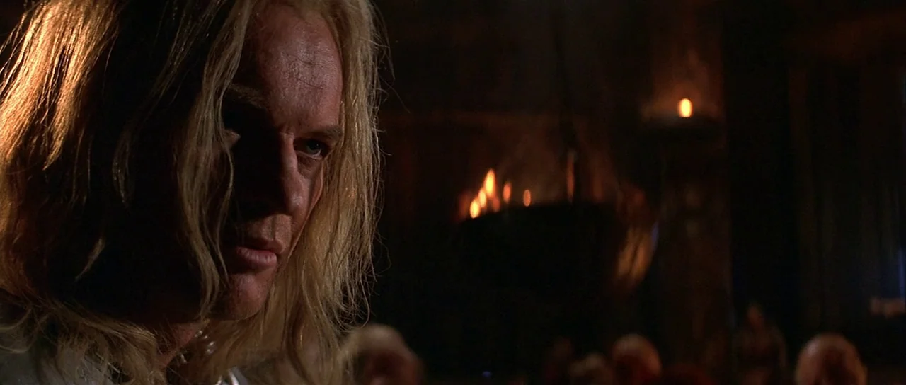The 13th Warrior (1999) movie still: closeup — A dark, expansive stone hall features a large, flickering fire in the background. A man with long…