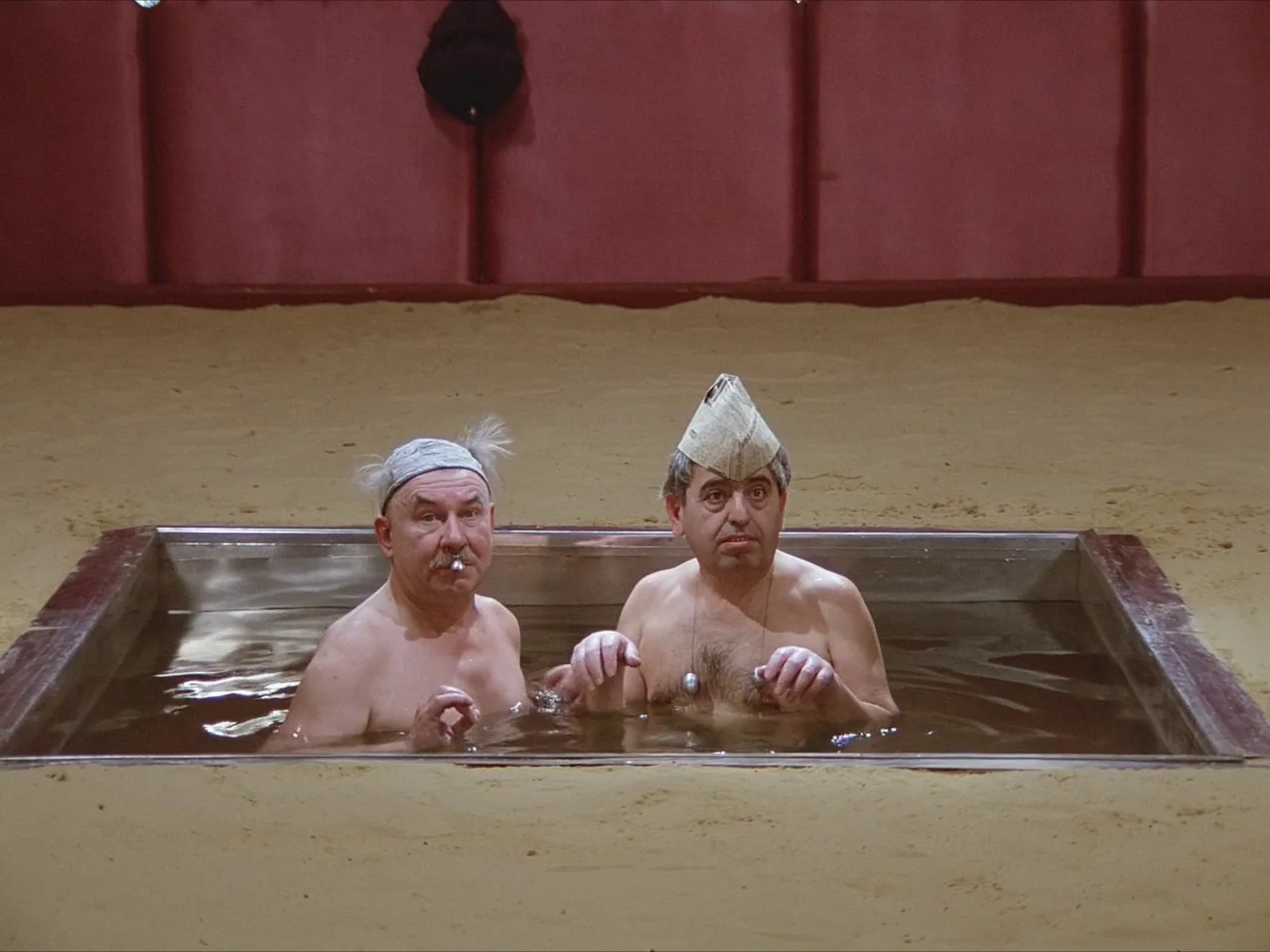 Kin-dza-dza! (1986) movie still: medium — Two men sit submerged in a metal tank set into a floor covered with sand. The backdrop consists of large,…