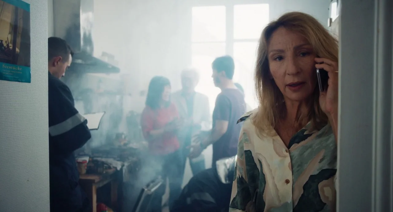 Anaïs in Love (2021) movie still: medium — A smoke-filled room contains several people standing in the background near a window. An older woman in…