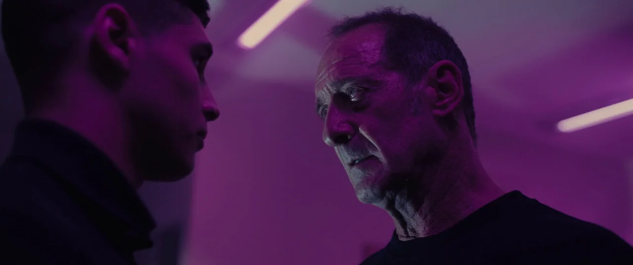 Titane (2021) movie still: medium — A dimly lit interior space with rows of linear purple light fixtures on ceiling. Two men stand facing each other…