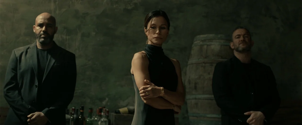 Hounds of War (2024) movie still: medium — A dark, textured stone wine cellar provides the backdrop for three individuals. One woman stands between…