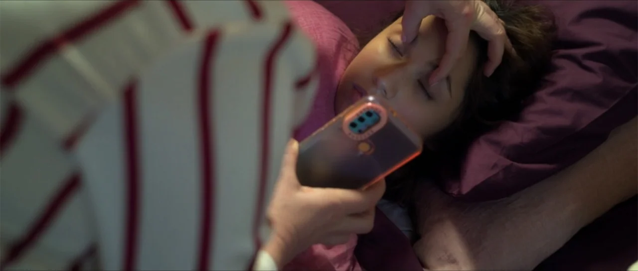 Disconnected 2 (2025) movie still: closeup — A dimly lit bedroom features purple bedding and a person lying with eyes closed. A second person in a…