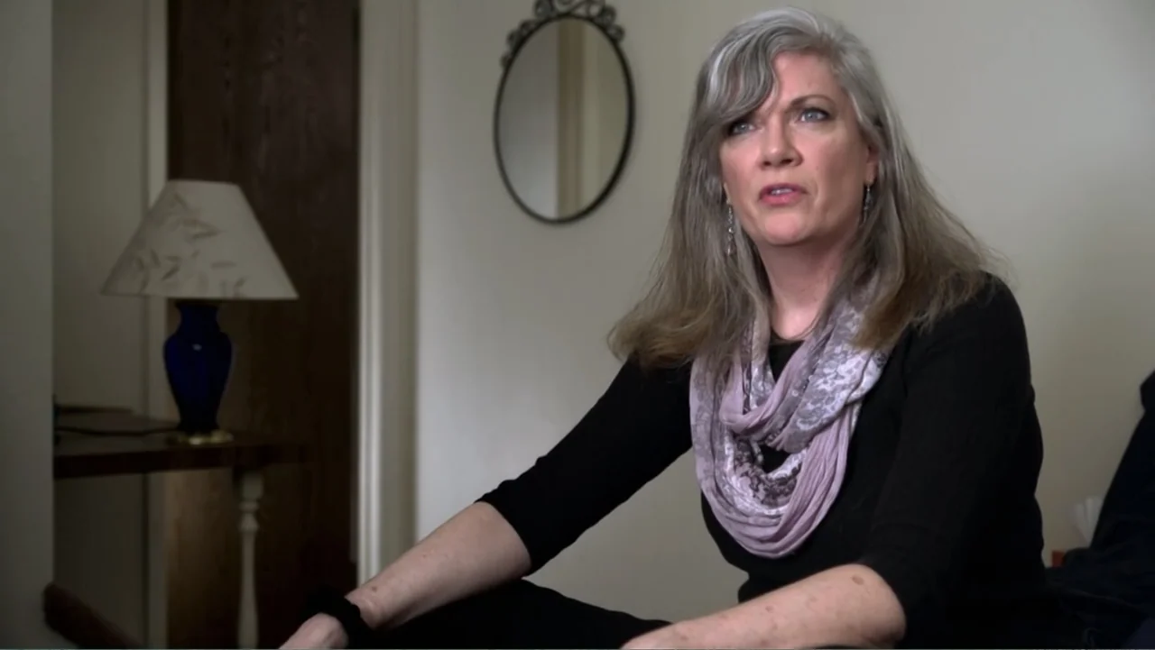 A Dangerous Son (2018) movie still: medium — A residential room features a plain wall with a small decorative oval mirror. A woman with grey hair…
