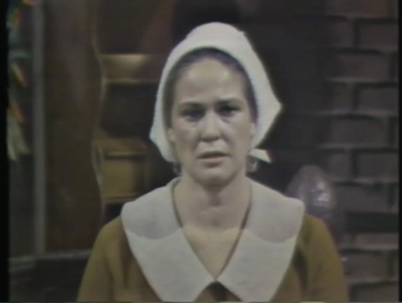 The Crucible (1996) movie still: medium — A dimly lit room with a dark brick wall visible in the background. A woman in a white head covering and…