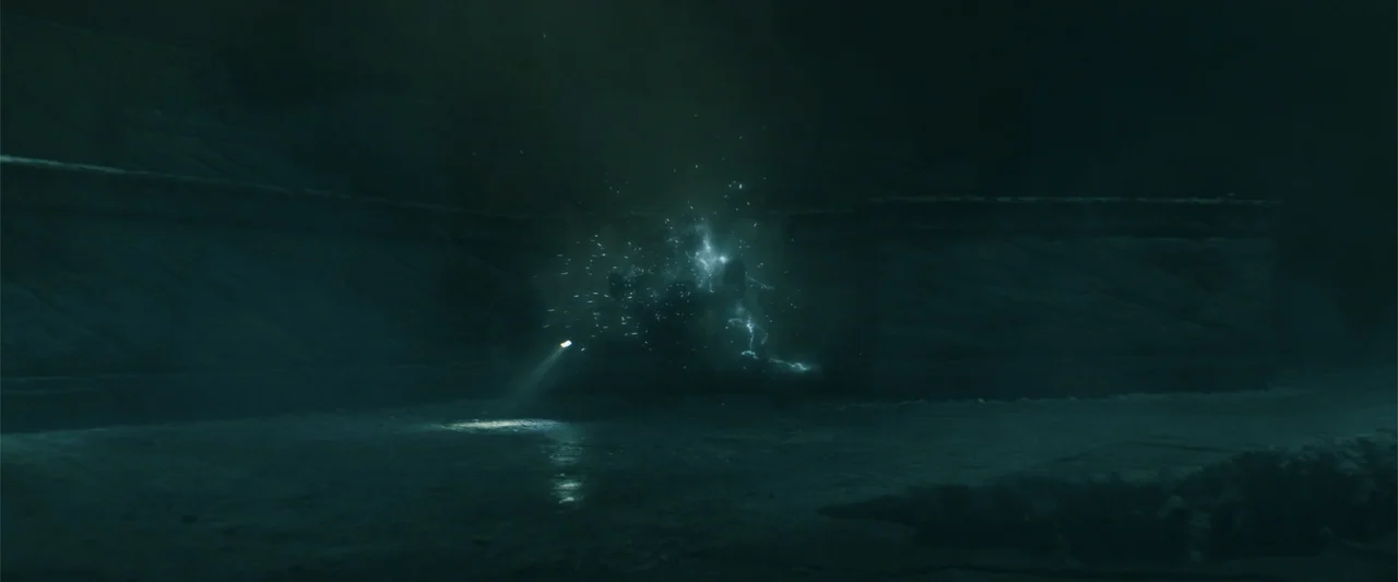 Black Adam (2022) movie still: wide — A dark subterranean stone chamber features rough-hewn walls and a flat floor. A localized burst of blue…