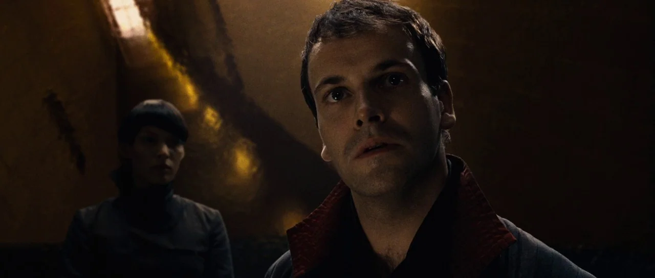 Aeon Flux (2005) movie still: medium — A dimly lit narrow space features textured dark walls with gold-toned highlights. Two people stand in the…