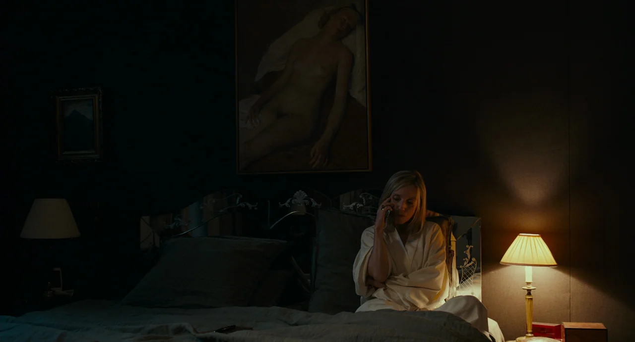 Last Summer (2023) movie still: medium — A dark bedroom with dark wallpaper contains a bed and a wall painting. A woman in a light-colored robe sits…