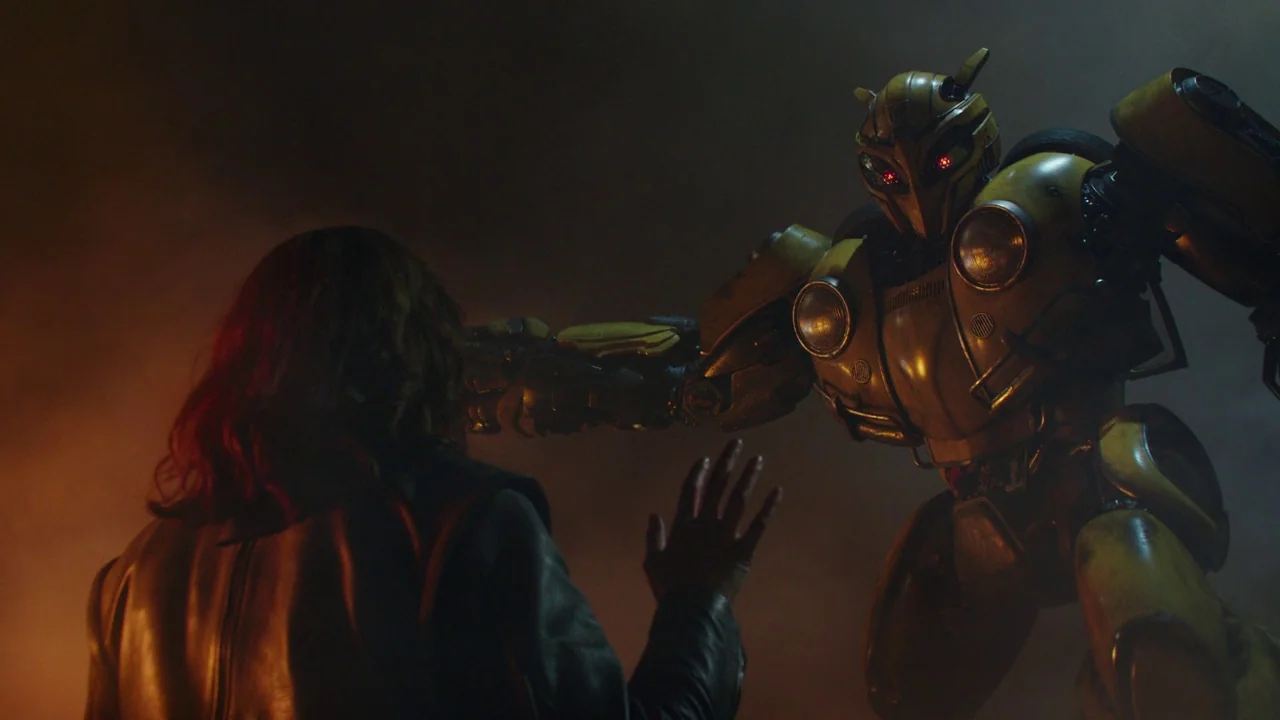 Bumblebee (2018) movie still: medium — A dark, smoky industrial wasteland at night illuminated by flickering warm light. A human woman reaches…