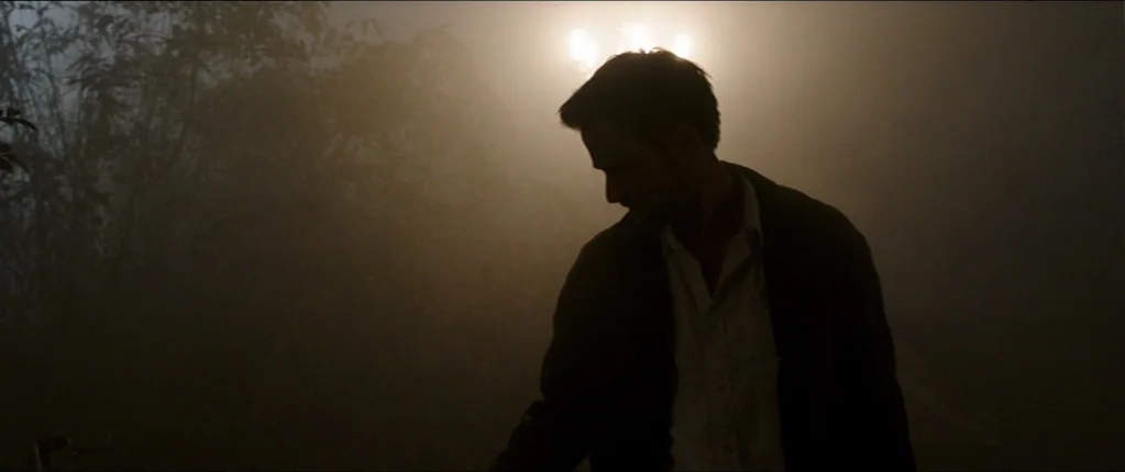 Abattoir (2016) movie still: medium — A dark city street is enveloped in a thick, low-hanging fog at night. A man stands in front of a glowing…