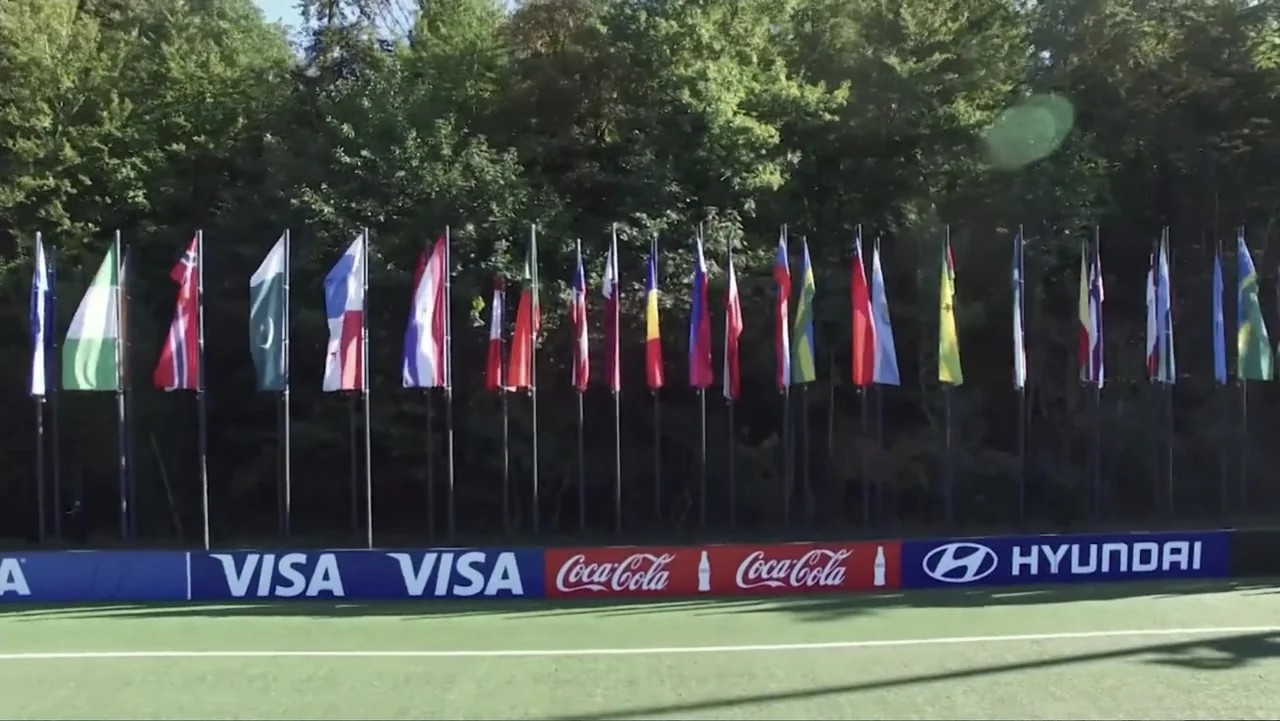 The FIFA Family: A Love Story (2017) movie still: wide — A row of various national flags stands on poles before a dense treeline. A long sponsor…