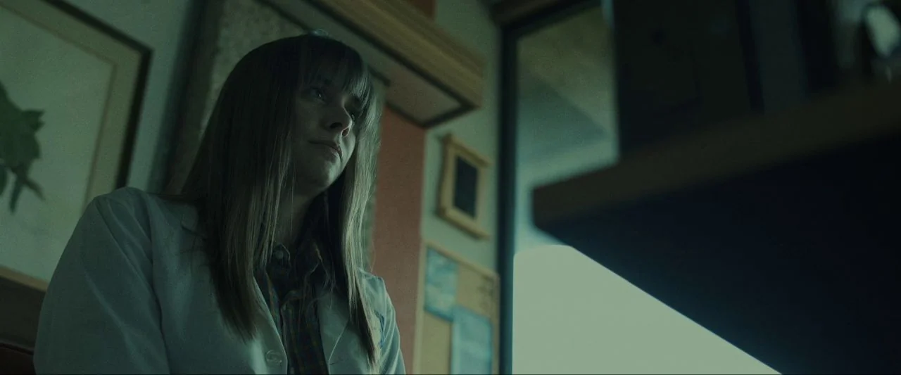 Come True (2020) movie still: medium — A professional interior space features a cluttered bulletin board and framed art on walls. A woman in a white…