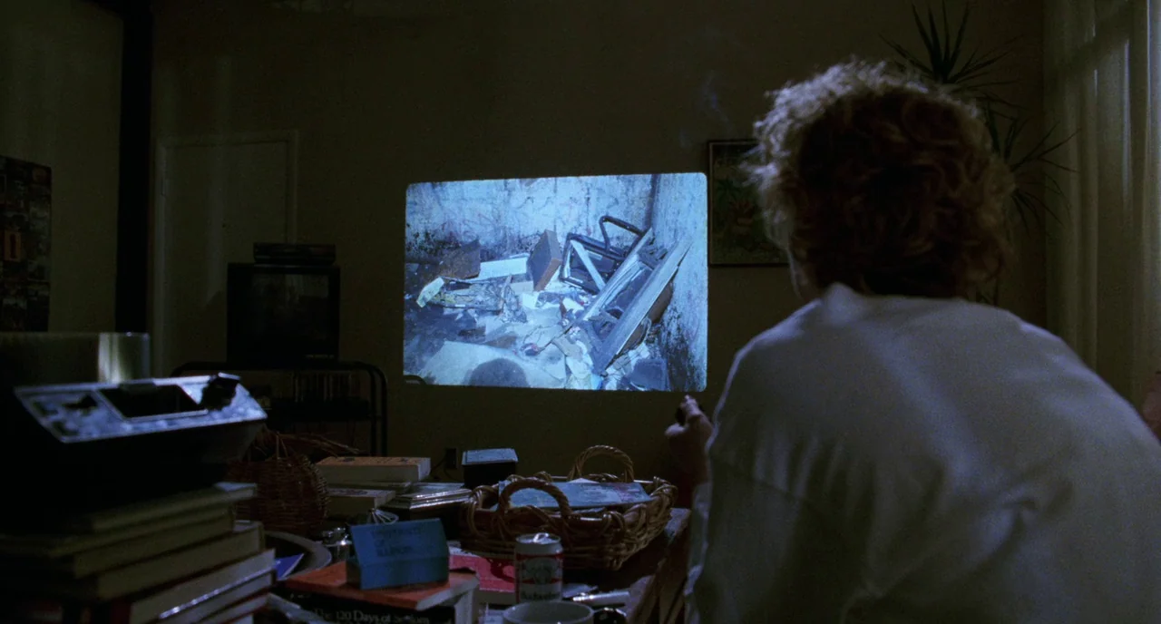 Candyman (1992) movie still: medium — A dark, cluttered interior room with stacks of books and a basket on a table. A person sits watching a film…