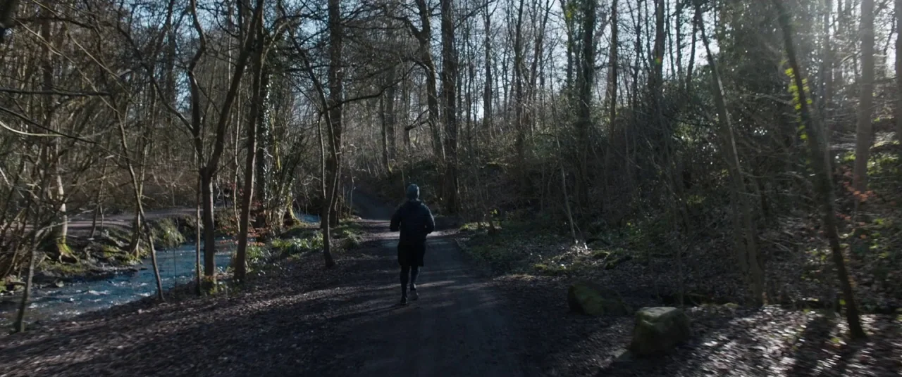 Journeyman (2017) movie still: wide — A narrow dirt trail winds through a leafless forest beside a shallow flowing stream. A person in dark athletic…