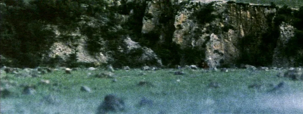 Japón (2002) movie still: establishing — A rocky, uneven terrain sits at the base of a textured limestone cliff. Sparse vegetation grows in the…
