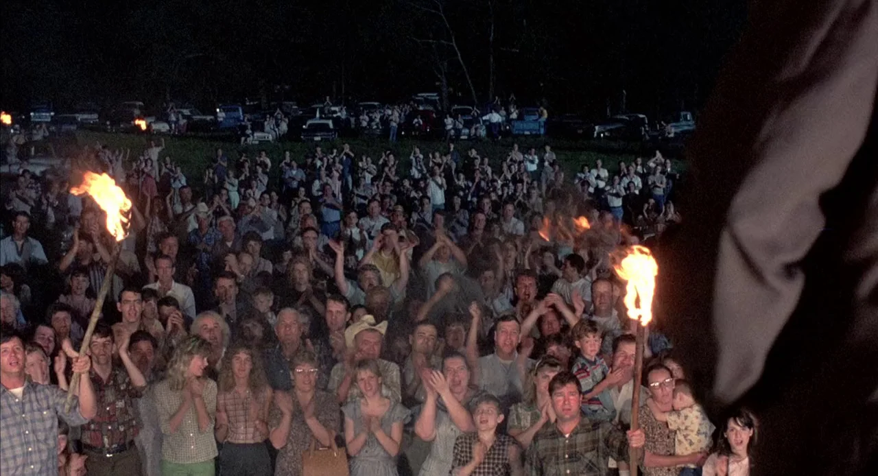 Mississippi Burning (1988) movie still: wide — A dense crowd of people stands at night in a grassy outdoor area. Individuals are clapping and…