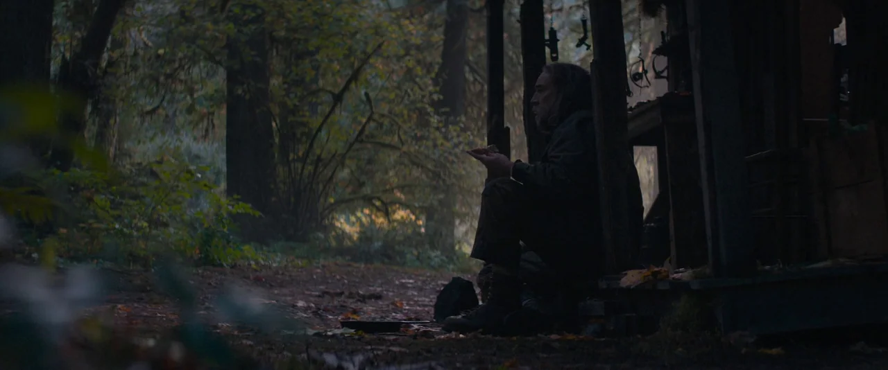 Pig (2021) movie still: medium — A man sits on the porch of a rustic wooden structure surrounded by dense trees. He is eating while positioned in…
