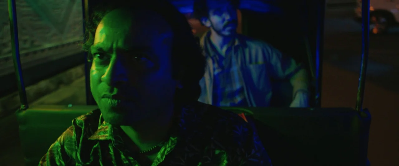 Monkey Man (2024) movie still: medium — Interior of a moving auto rickshaw at night with dark exterior surroundings. Two men sit close together…