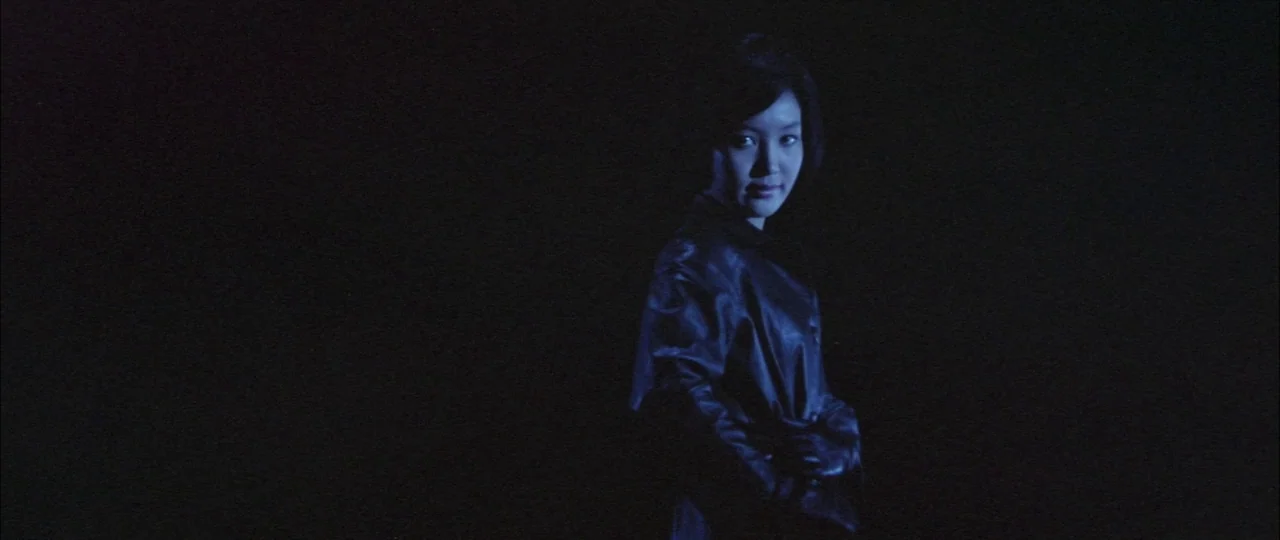 Black Tight Killers (1966) movie still: medium — A dark space with no visible architecture or environmental details. A woman in a shiny coat looks…