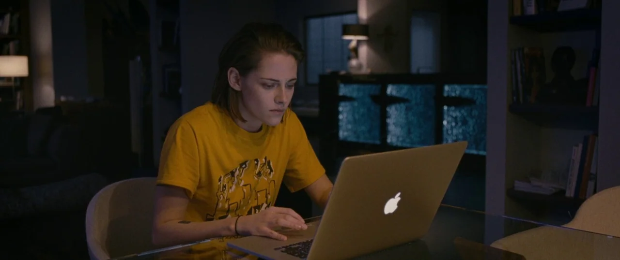 Personal Shopper (2016) movie still: medium — A modern home interior with glass surfaces and recessed shelving in the background. A woman sits at a…