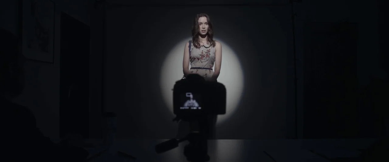 Starry Eyes (2014) movie still: medium — A dark, spartan studio space features a centered subject within a harsh circular beam. A digital camera on…