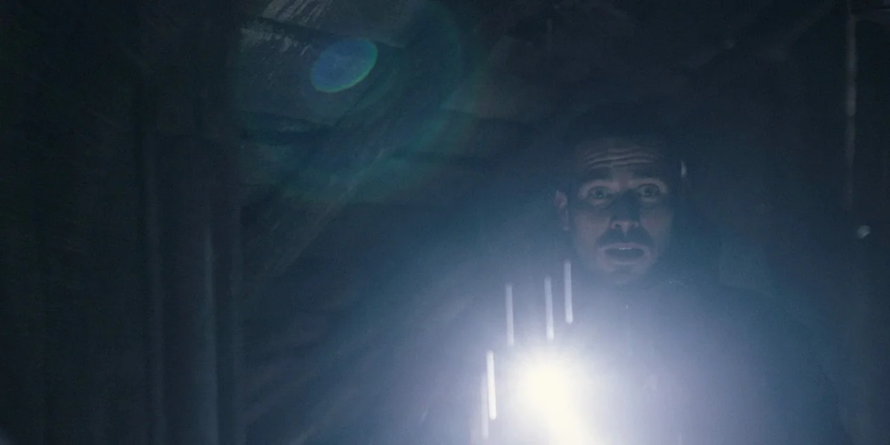 The Ritual (2017) movie still: closeup — A dark, cramped space with rough wooden rafters visible in the background. A man shines a flashlight…
