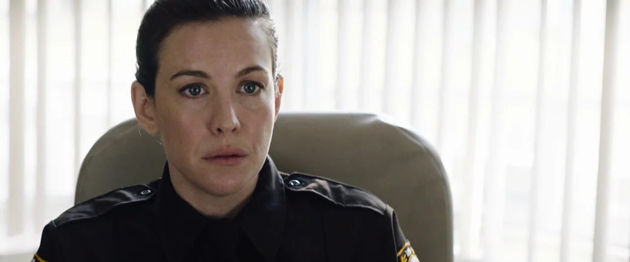 Wildling (2018) movie still: closeup — An interior space with bright light coming through vertical window blinds. A woman in a dark police uniform…