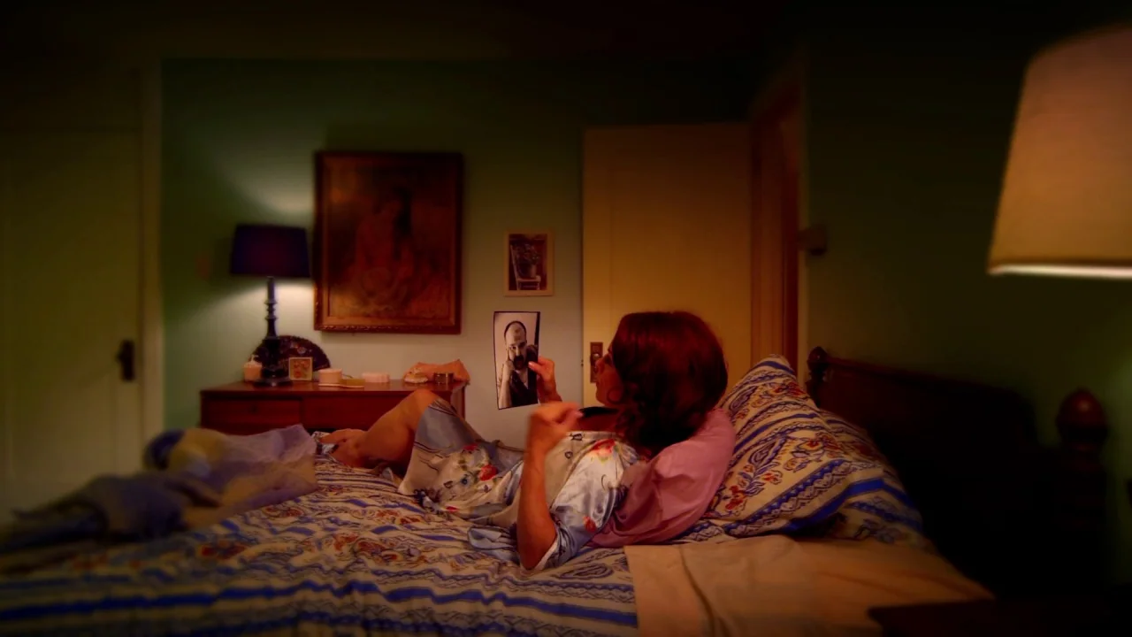 Suburban Gothic (2014) movie still: medium — A dimly lit bedroom features a bed with patterned linens and an old painting. A woman reclines against…
