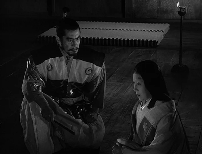 Throne of Blood (1957) movie still: medium — A dark, wood-floored room features a low mattress on the floor in the background. A samurai in a…