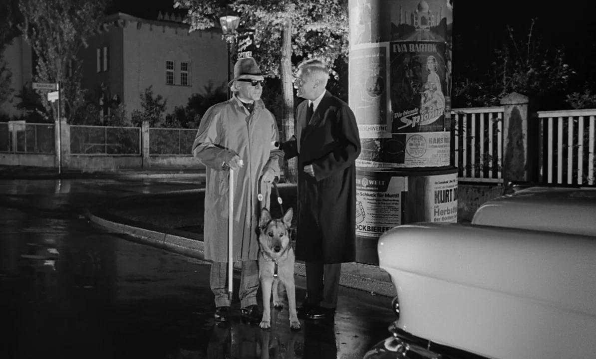 The 1,000 Eyes of Dr. Mabuse (1960) movie still: fullbody — A nighttime city street corner features a cylindrical poster column covered in…