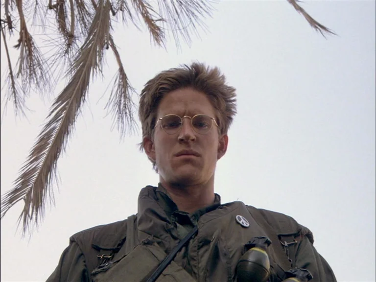 Full Metal Jacket (1987) movie still: medium — A soldier stands amidst tropical palm fronds against an overcast pale sky. The man in military gear…