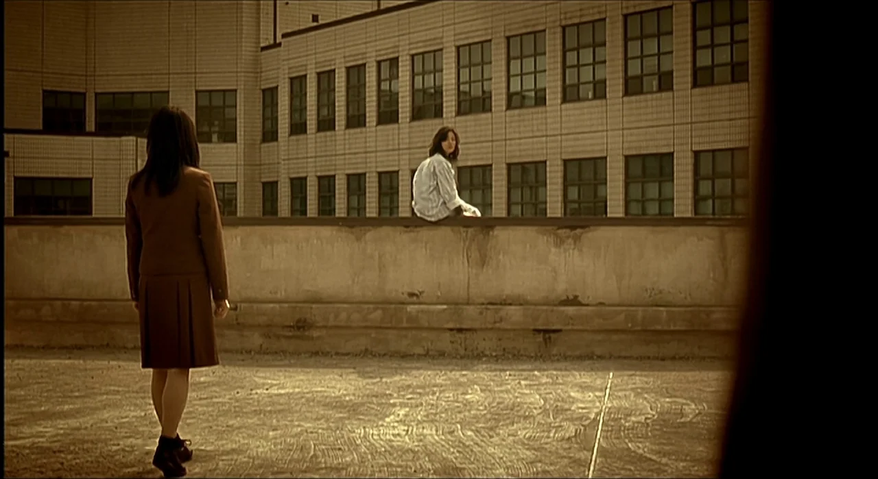 Voice (2005) movie still: wide — A flat rooftop terrace extends toward a large, multi-story tiled school building. One girl walks across the…