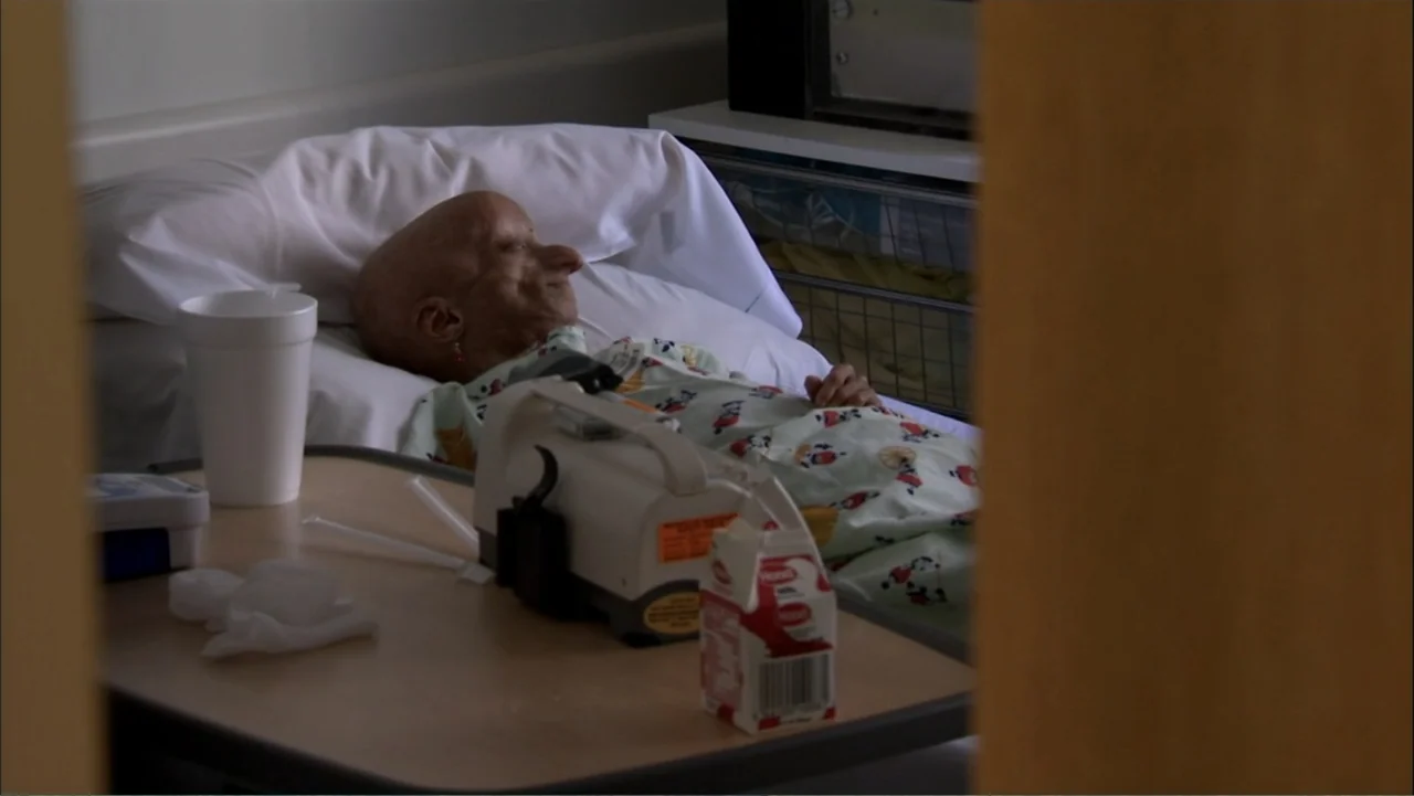 Life According to Sam (2013) movie still: medium — A sterile hospital room contains a bed with white pillows and a patient. An elderly woman in a…