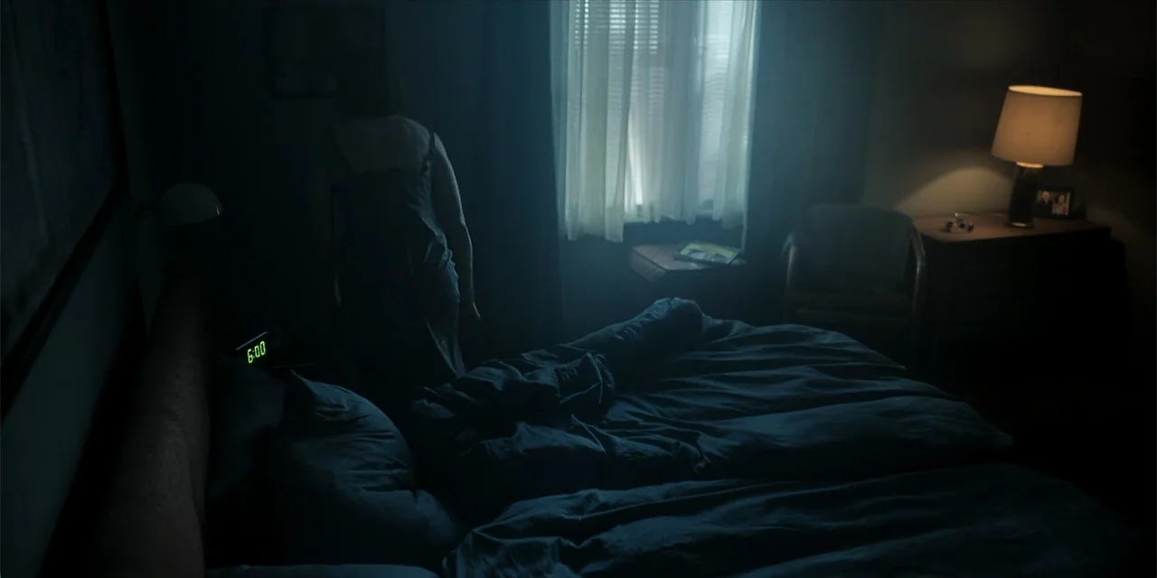 Oslo (2021) movie still: medium — A dimly lit room contains a rumpled bed and a table lamp on a nightstand. A woman in a slip dress stands with her…