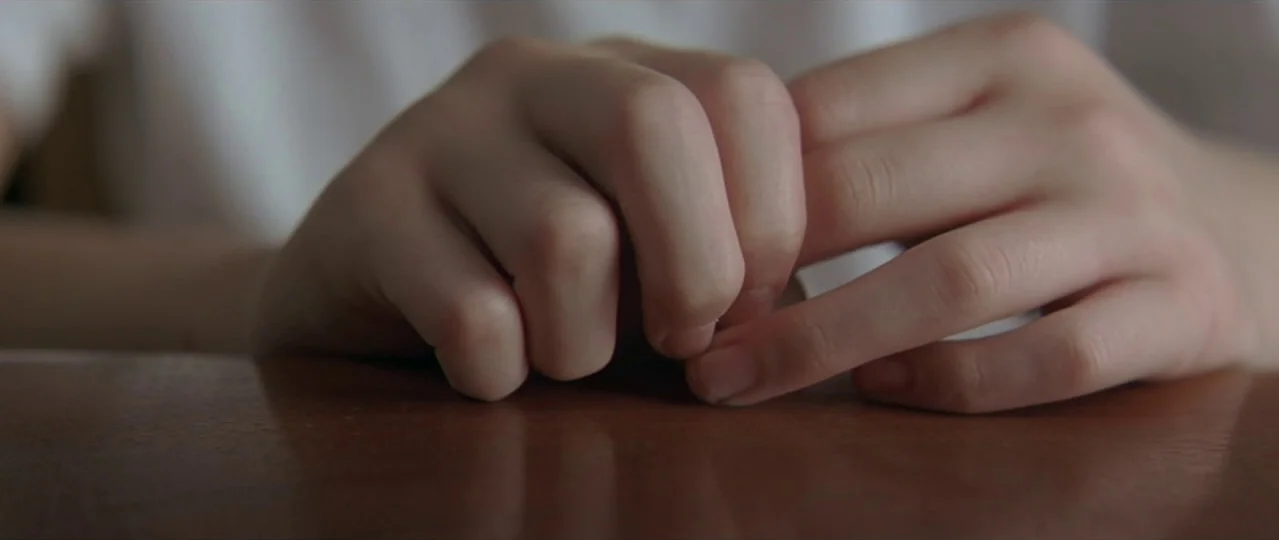 Afterschool (2008) movie still: closeup — A pair of hands rests on a smooth, dark-stained wooden table surface. The hands are positioned together as…