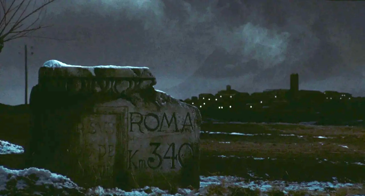 Roma (1972) movie still: wide — A dark rural landscape with a weathered stone milestone in the foreground. In the distance a small town sits on a…