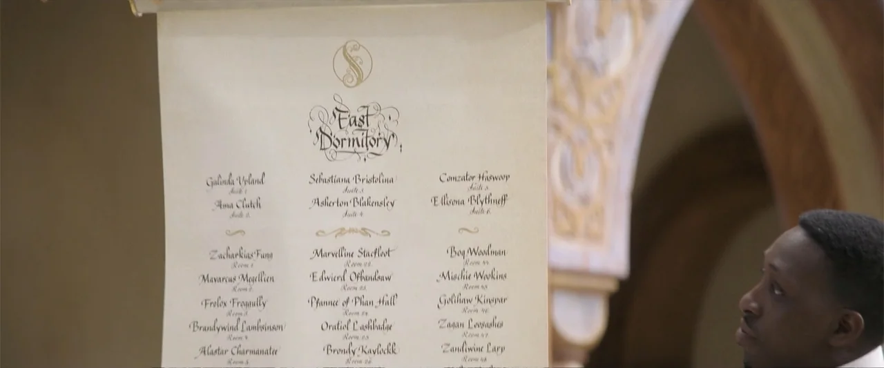 Wicked (2024) movie still: closeup — An interior space with a hanging parchment scroll detailing room assignments at East Dormitory. A man stands at…