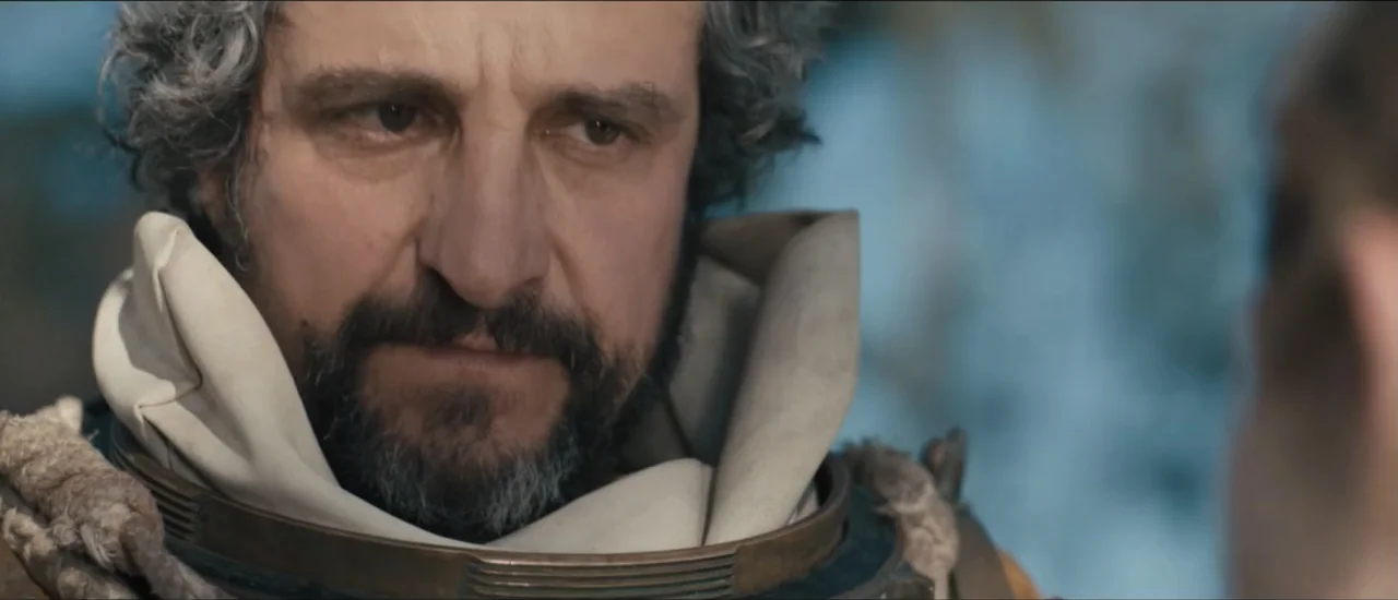 Fear Below (2025) movie still: closeup — A man with a graying beard wears a metallic collar and fabric garment outdoors. He maintains a steady gaze…