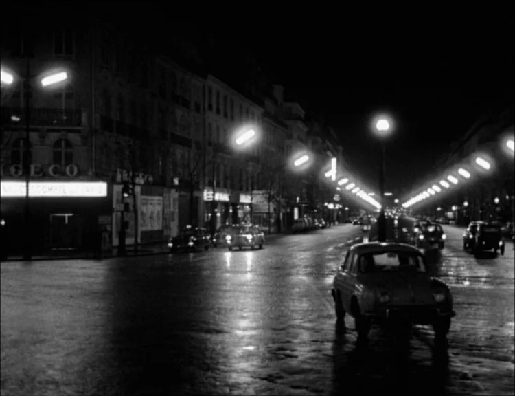 Alphaville (1965) movie still: establishing — A wide view of a Parisian city street at night with wet asphalt. Multiple cars drive along the road…